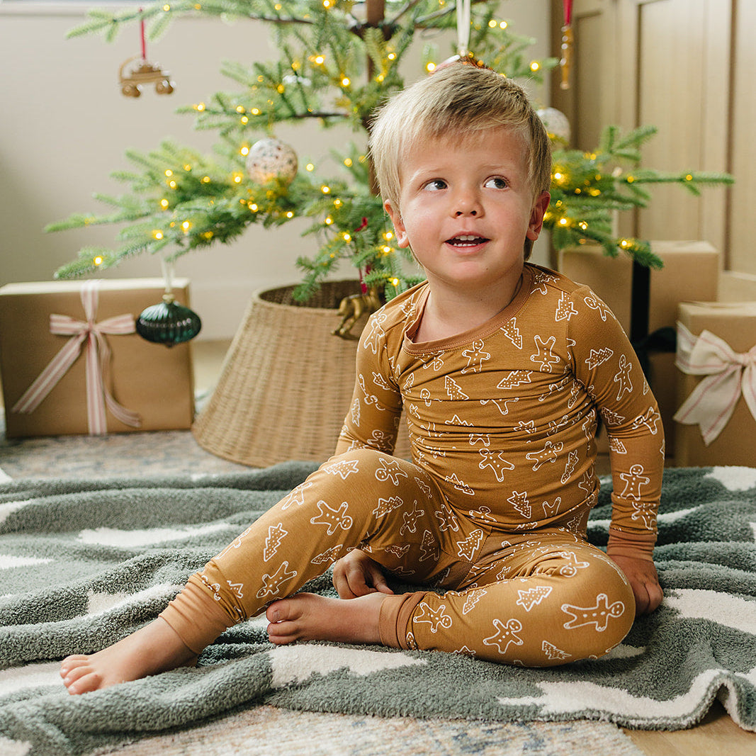 Child in a mustard yellow onesie sitting on a blanket in front of a decorated Christmas tree.