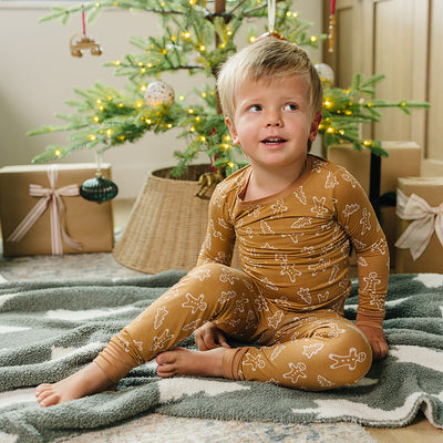 Child in a mustard yellow onesie sitting on a blanket in front of a decorated Christmas tree.