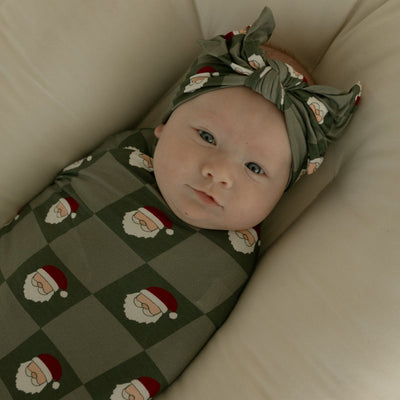 Baby wrapped in a green blanket with Santa Claus patterns, wearing a matching headband, lying on a beige surface.