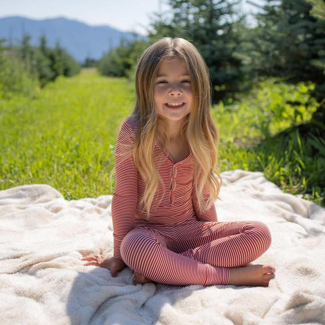 A young girl with long blonde hair sits cross-legged on a cream blanket in a sunny outdoor field, wearing a pink ribbed long-sleeve pajama set. Trees and mountains are visible in the background.