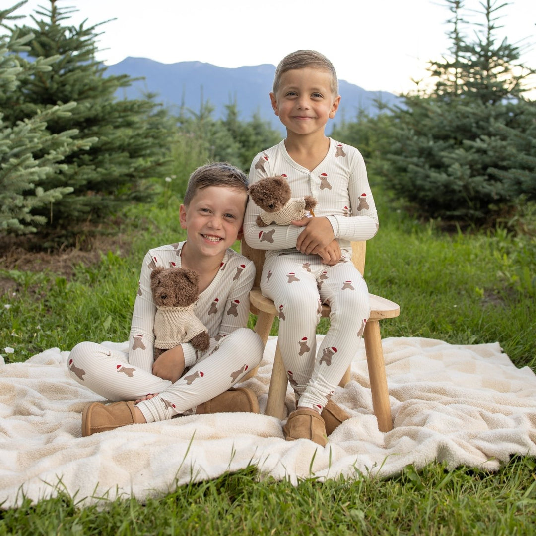 Two children wearing matching cream-colored pajamas with a brown bear print, sitting on a white blanket on a grassy lawn with trees and mountains in the background.