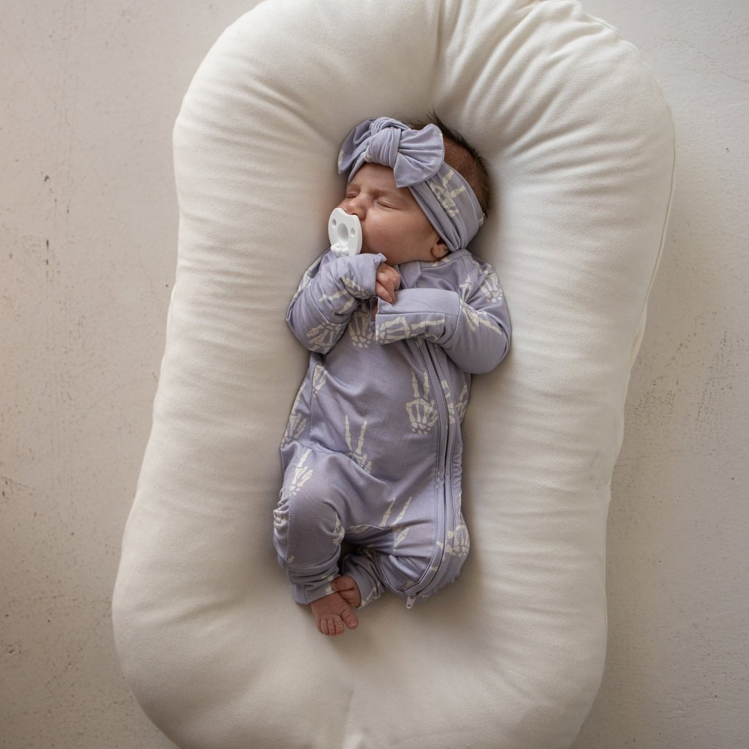 A sleeping newborn baby wearing a light lavender-blue patterned onesie and matching headband, lying on a large cream-colored U-shaped baby support pillow against a neutral beige wall.