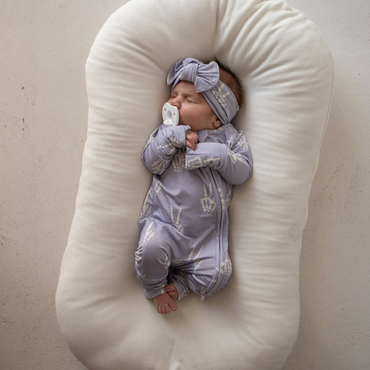 A sleeping newborn baby wearing a light lavender-blue patterned onesie and matching headband, lying on a large cream-colored U-shaped baby support pillow against a neutral beige wall.
