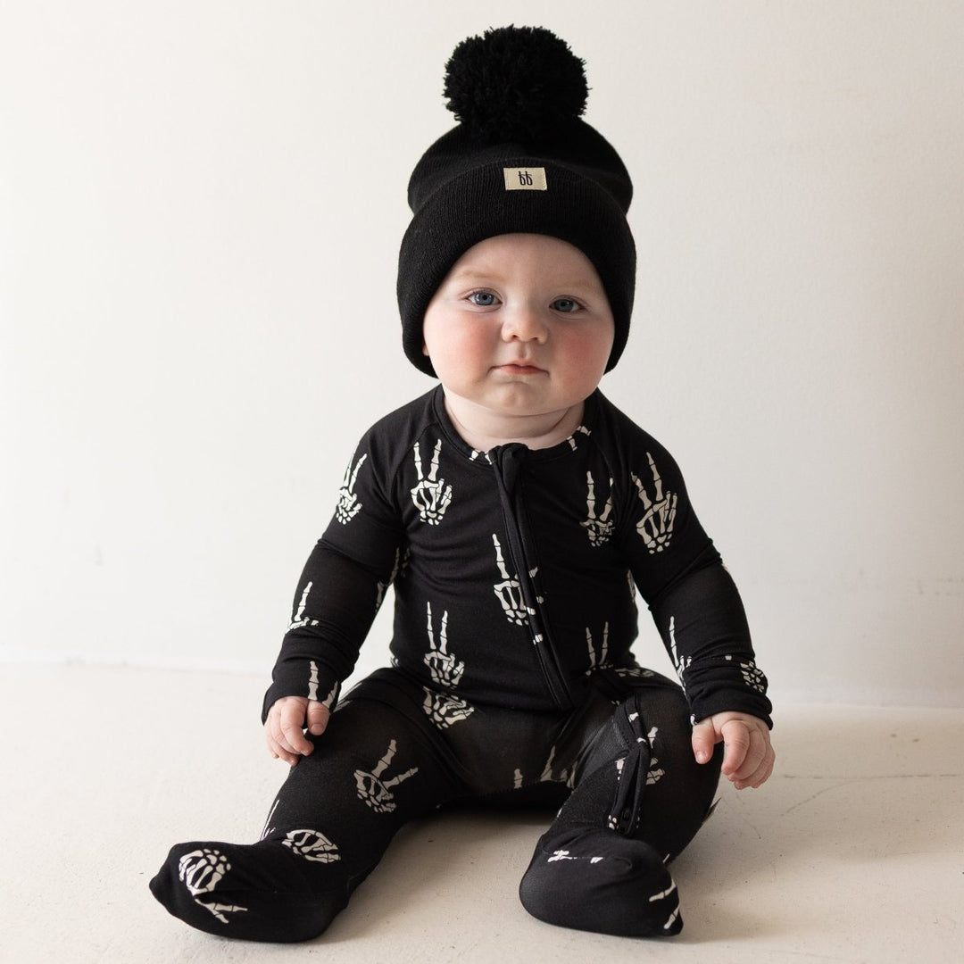A baby sitting on a light beige floor wearing a black knit beanie with a pom-pom and a black footed sleeper with white skeleton handprints.