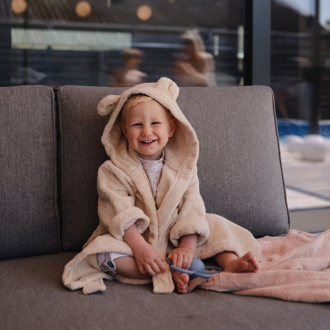 Child in a beige robe with a hood sitting on a gray couch.