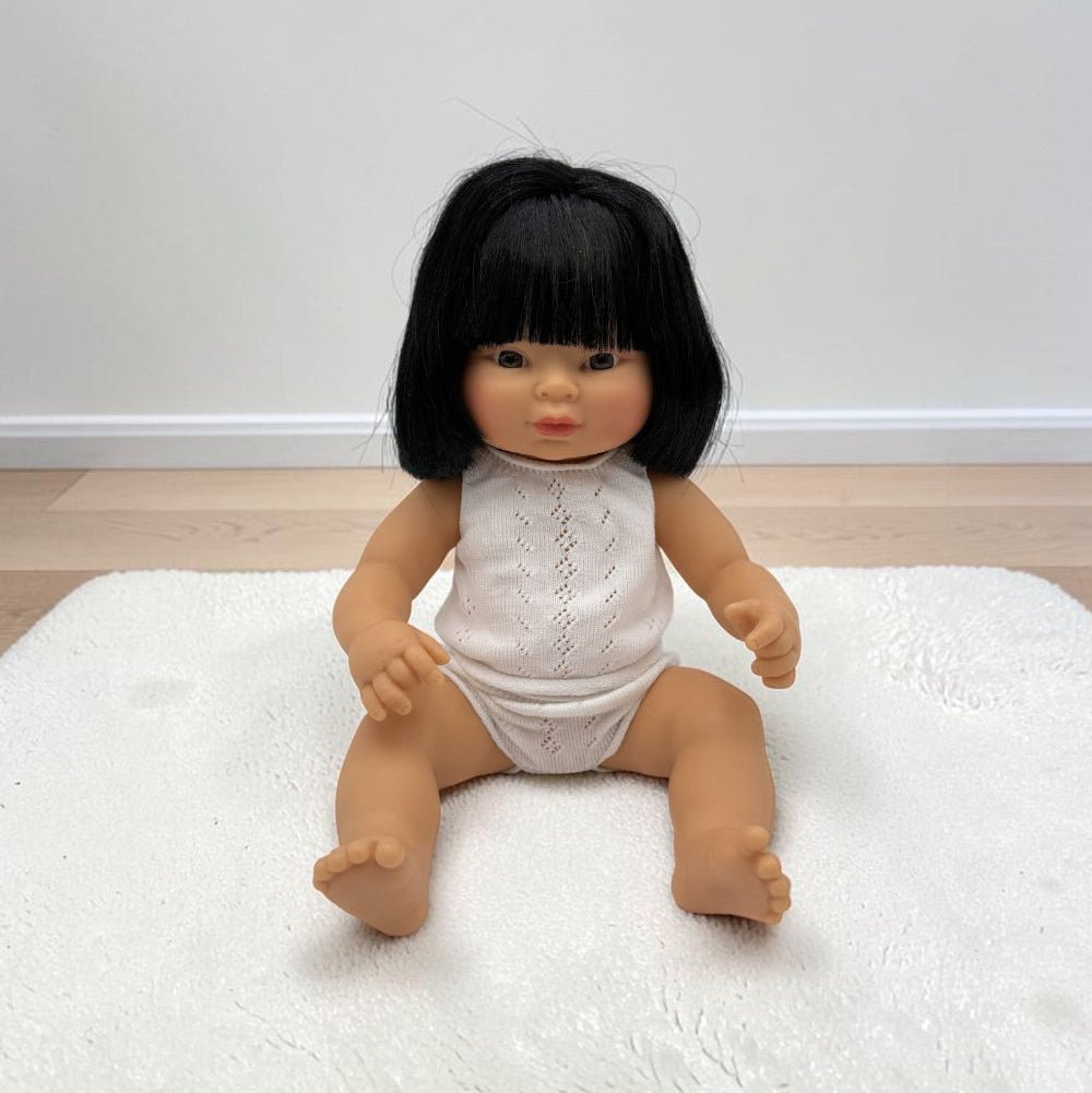A realistic Asian baby girl doll with black hair sitting on a white textured rug in front of a plain wall.