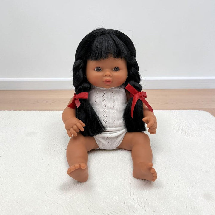 A realistic Hispanic baby doll with dark brown skin, black braided hair, and a white knit top with red ribbon accents, sitting on a white textured rug.