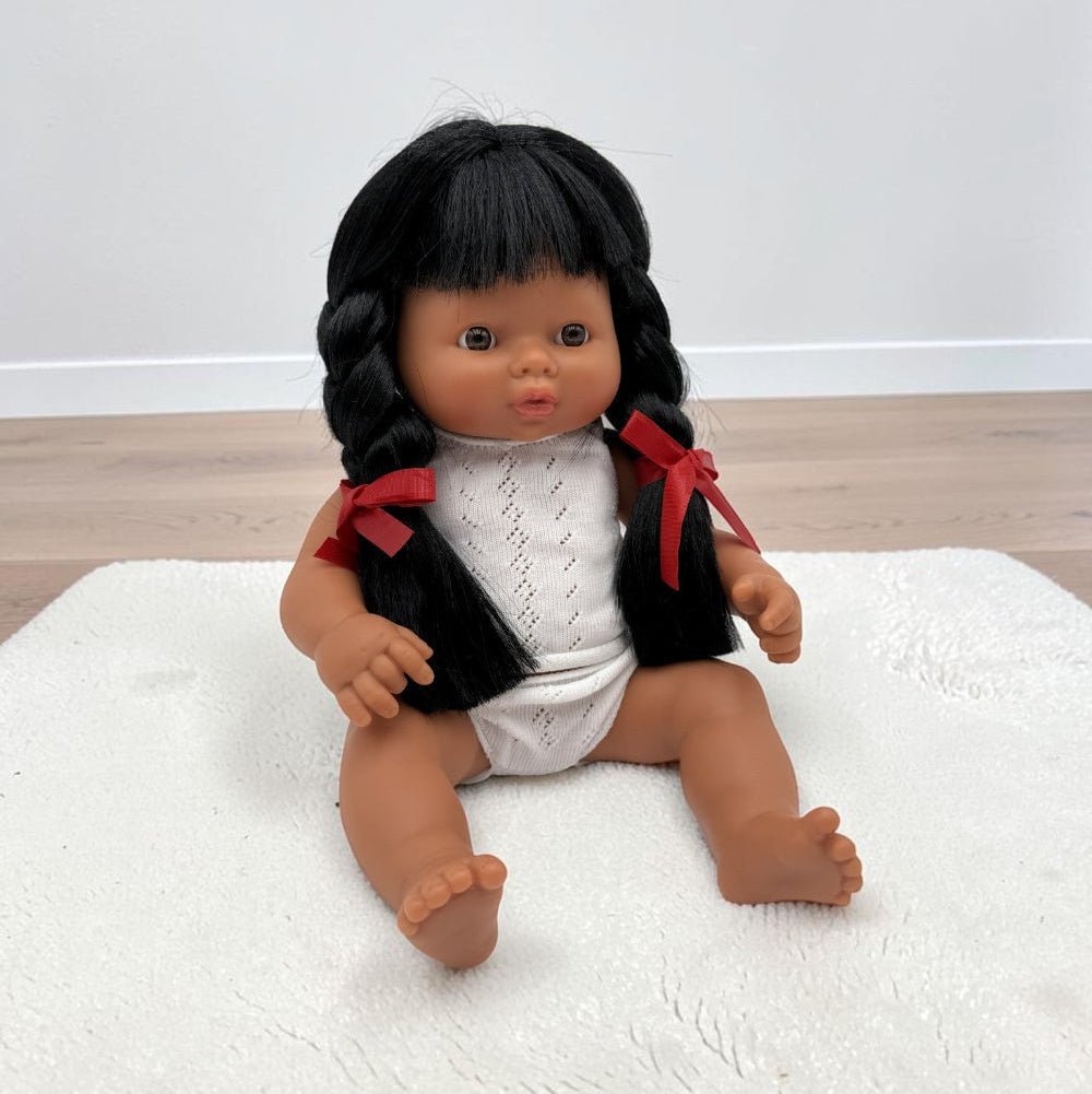 A Hispanic baby doll with black braided hair and red ribbon accents, wearing a white knit top and white diaper, sitting on a white textured rug.