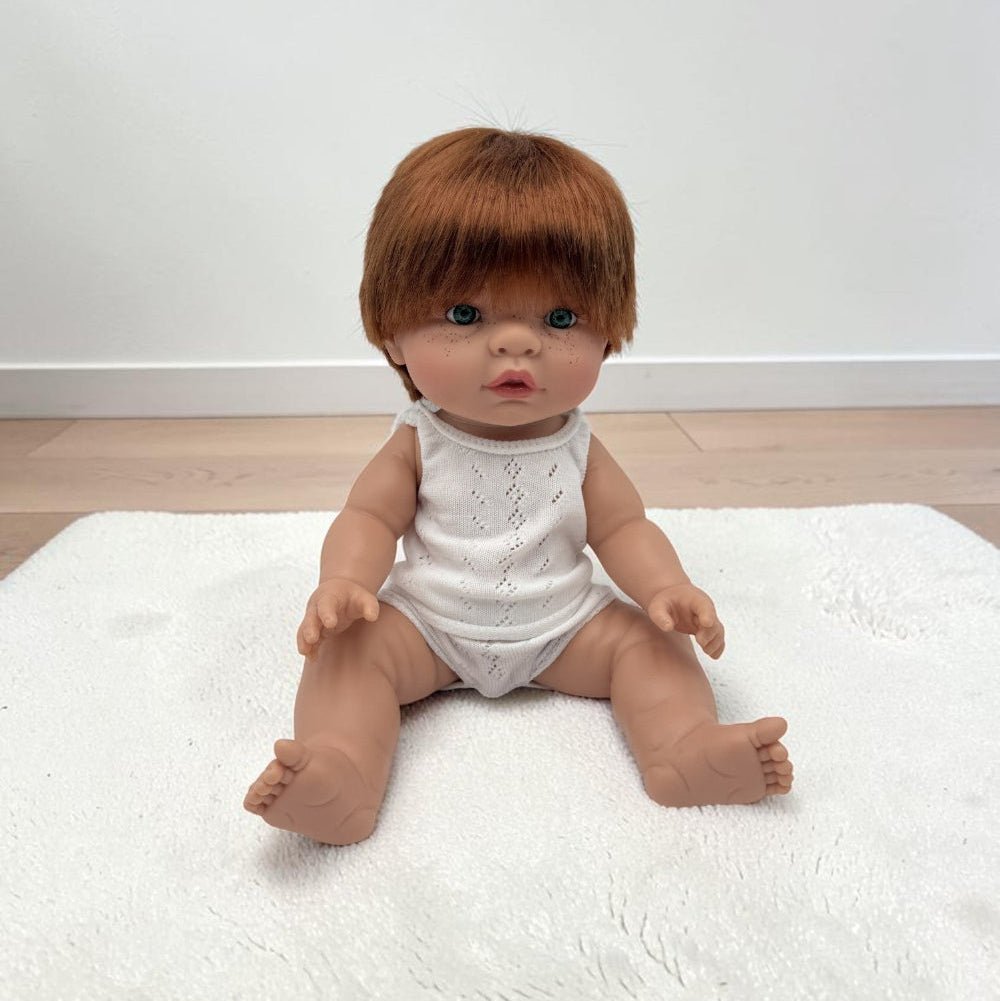 A realistic baby doll with short brown hair sits on a white textured rug against a plain white wall and light wood floor.