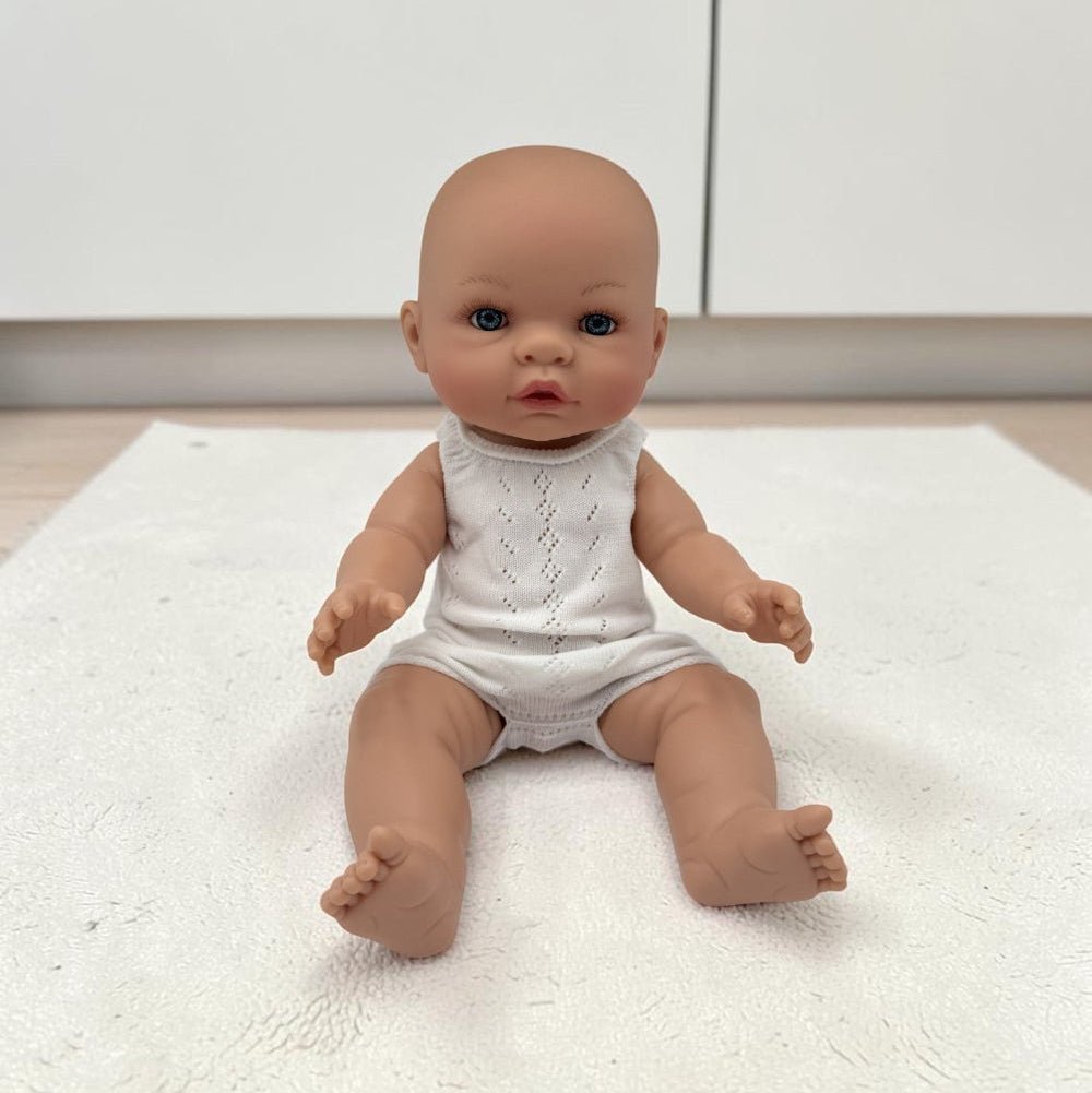 A realistic baby doll with a bald head and blue eyes, wearing a white sleeveless knit romper, sitting on a white textured rug in front of white cabinets.