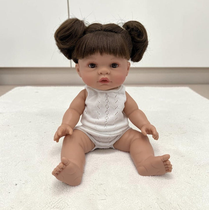 A realistic baby doll with brown hair styled in two buns, wearing a white sleeveless bodysuit, sitting on a white textured rug.