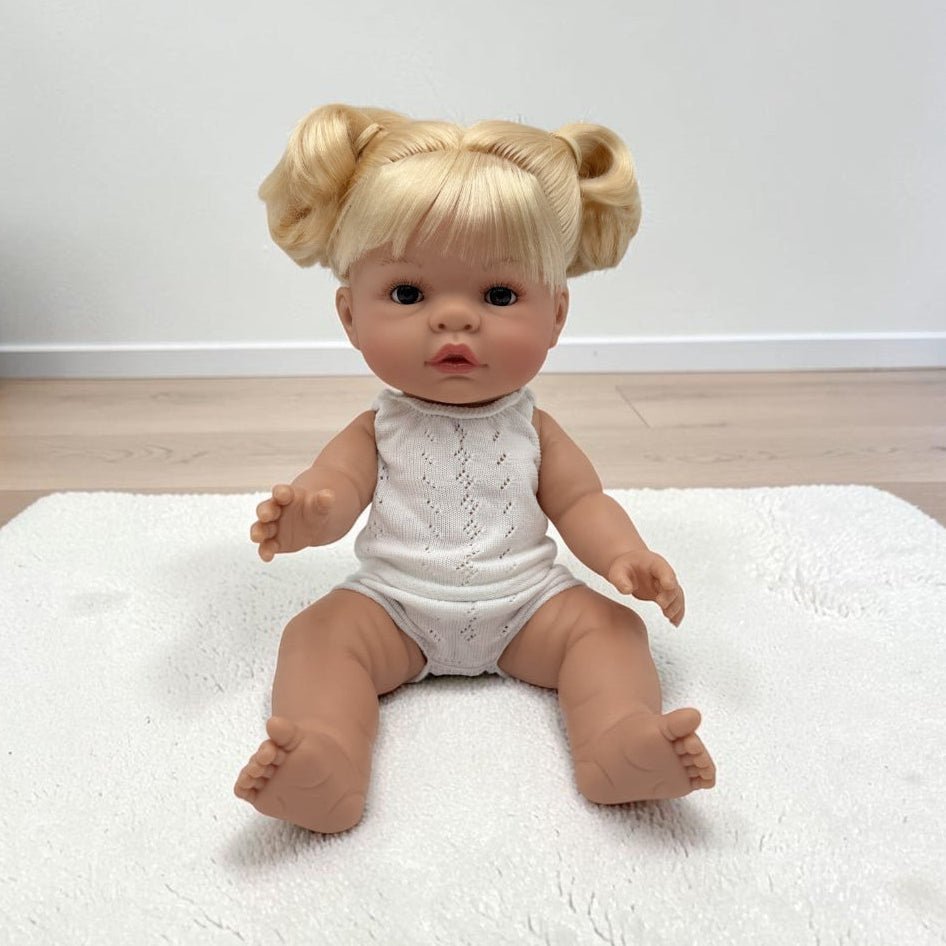 A realistic baby doll with blonde hair in two buns sits on a white textured rug in a room with light wood flooring and a white wall.