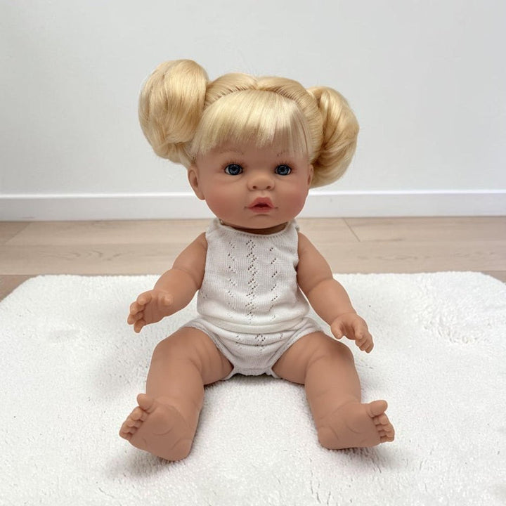 A realistic baby doll with blonde pigtails sits on a white textured rug in front of a plain white wall and light wood floor. The doll is wearing a white sleeveless knit romper.