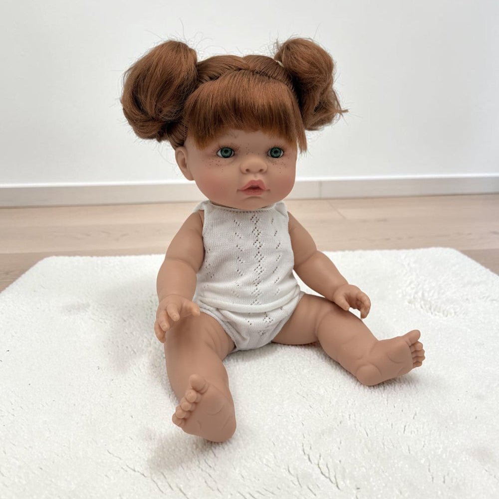 A realistic baby doll with red hair in two buns sits on a white textured rug. The doll has a realistic skin tone, blue eyes, and is wearing a white sleeveless knit romper.