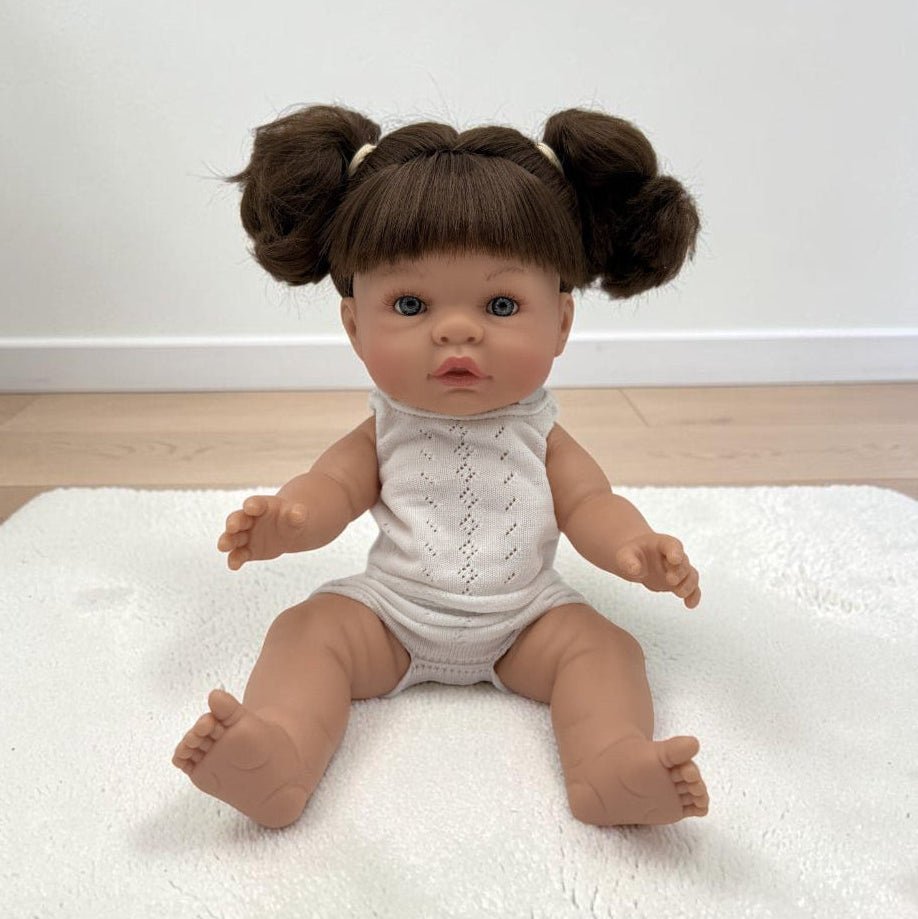 A realistic baby doll with brown hair in two buns sits on a white textured rug in a room with a white wall and light wood floor. The doll is wearing a white sleeveless knit bodysuit.
