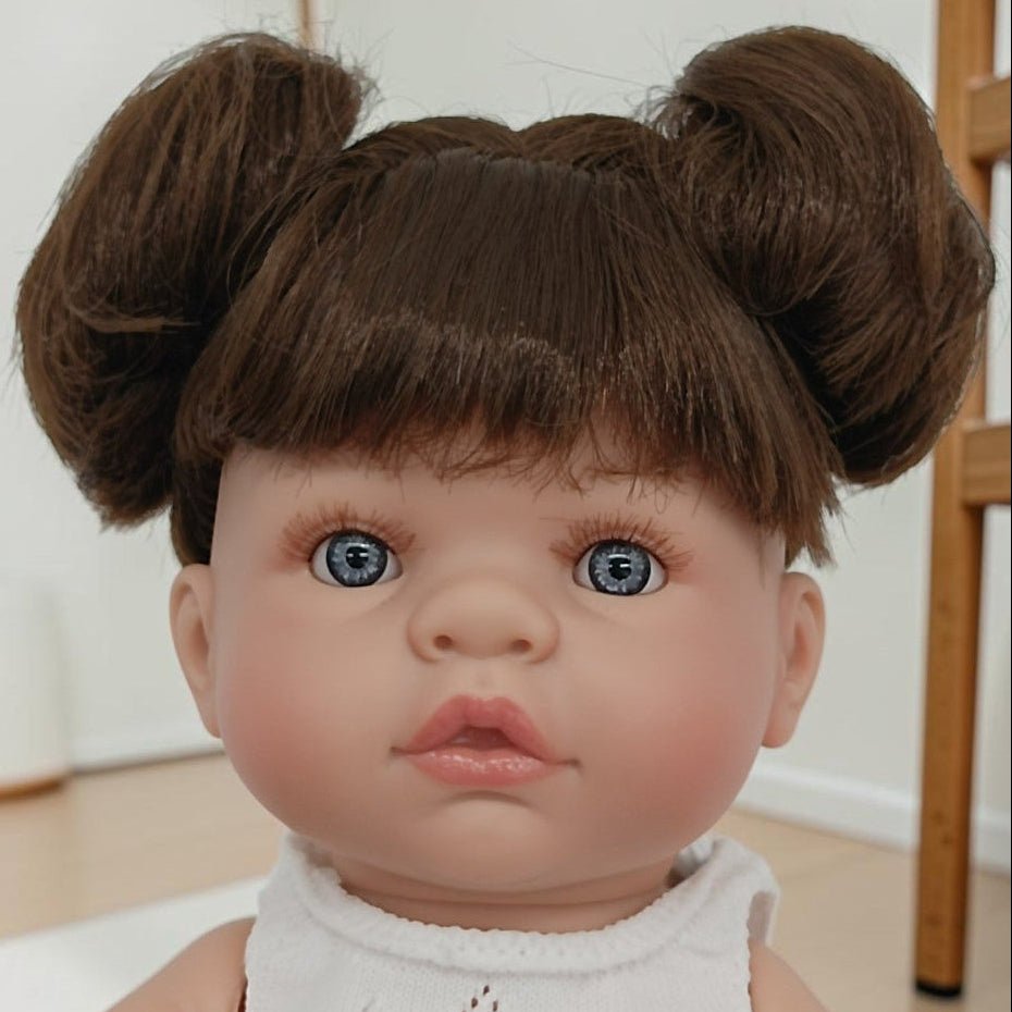 Realistic baby doll with brown hair styled in buns, wearing a white sleeveless top.