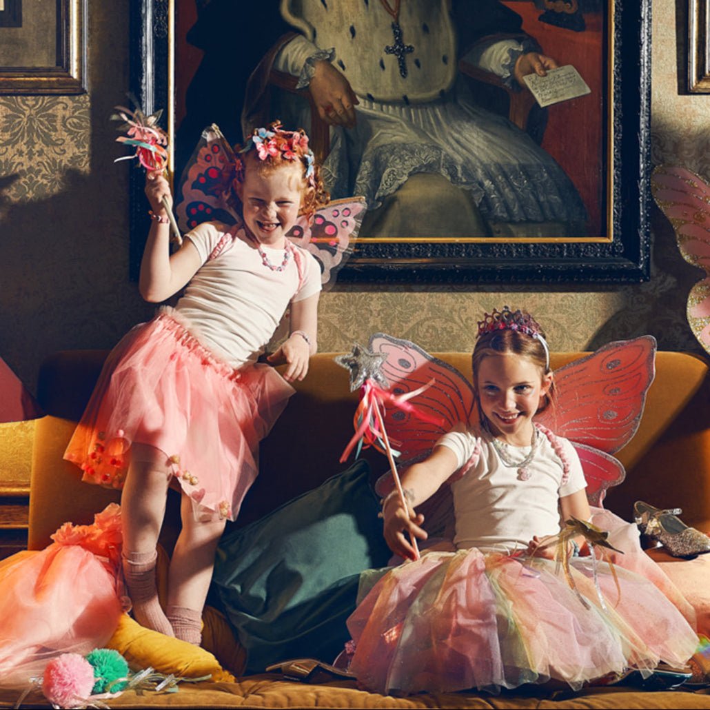 Two children in fairy costumes sitting on a couch with a classical painting in the background.