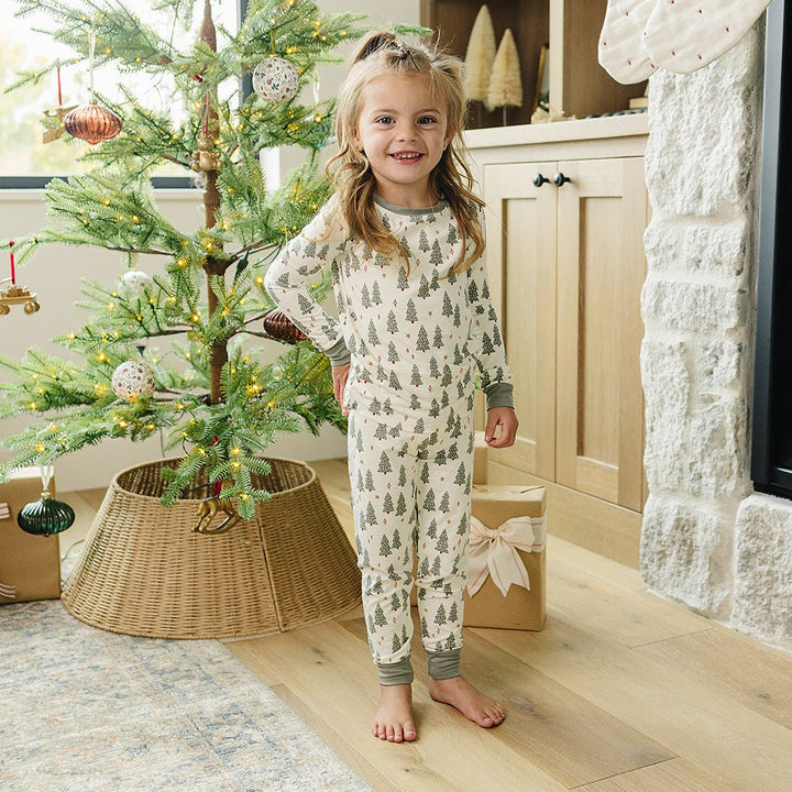Child in pajamas standing next to a decorated Christmas tree in a cozy living room.