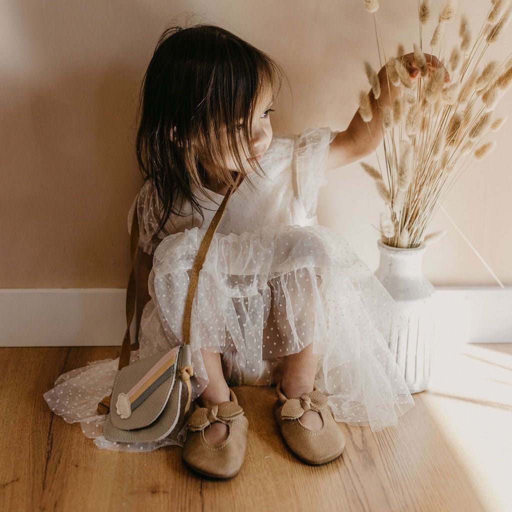 Young girl in a white dress standing next to a vase of dried flowers against a beige wall.