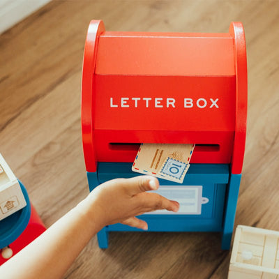 Red and blue toy letter box on a wooden floor with a hand placing a letter inside.