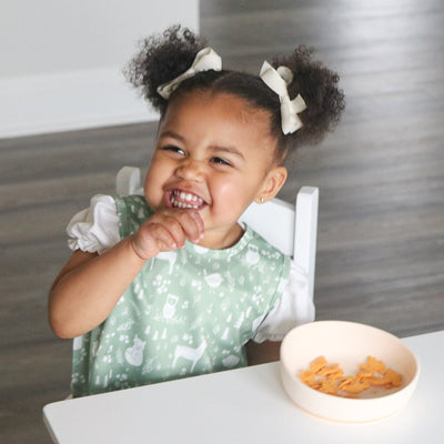 Child in a high chair with a bowl of food, wearing a green bib.