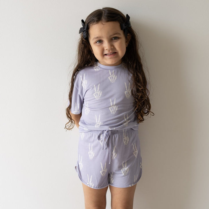 A young girl with long brown hair wearing a light purple short-sleeve t-shirt and matching shorts with a white peace sign pattern, standing against a plain off-white wall.