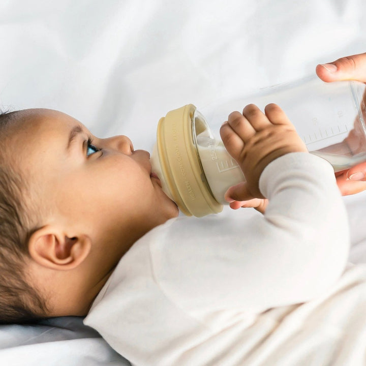 A baby in a white long-sleeve outfit drinking from a clear bottle with a beige cap, held by an adult hand against a soft white background.