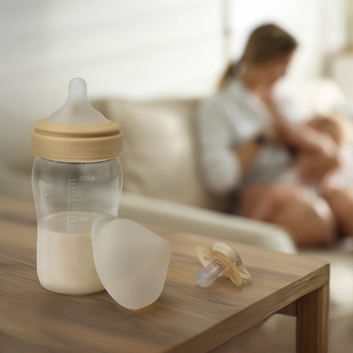 A baby bottle with a beige cap and nipple, a detached silicone breast shield, and a pacifier resting on a wooden table, with a softly blurred mother holding a baby in the background.