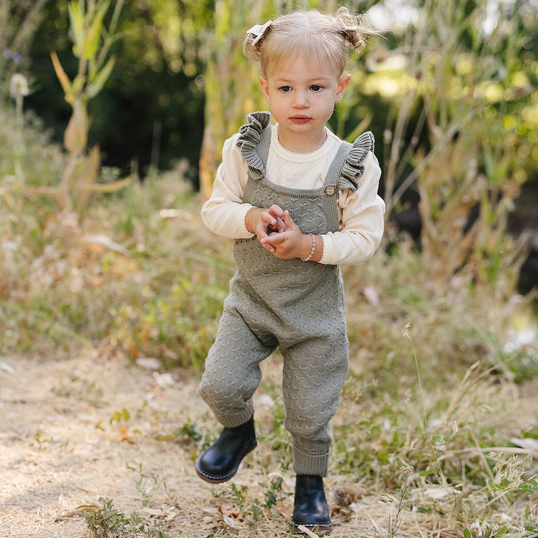 A child in a gray overall standing outdoors with plants in the background