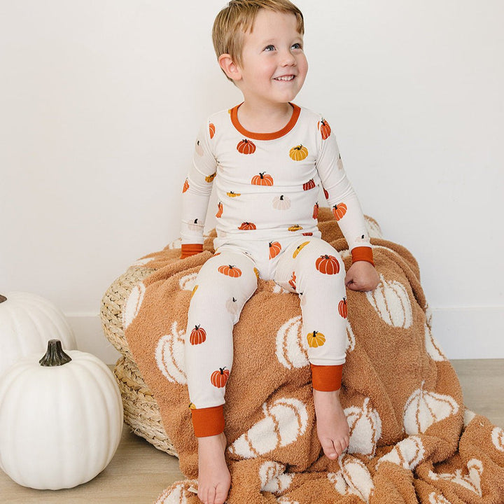 Child wearing pumpkin-patterned pajamas sitting on a plush blanket with pumpkins, next to a white pumpkin.