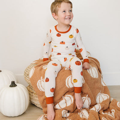 Child wearing pumpkin-patterned pajamas sitting on a plush blanket with pumpkins, next to a white pumpkin.