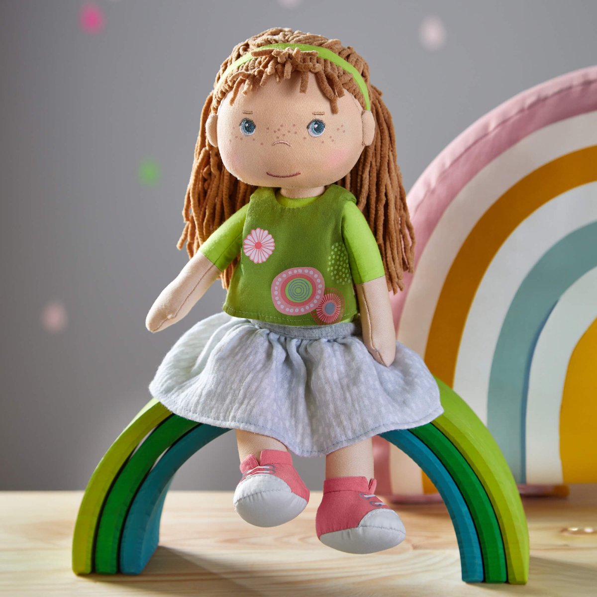 Doll with green top and white skirt standing on a wooden surface with a colorful rainbow toy in the background.