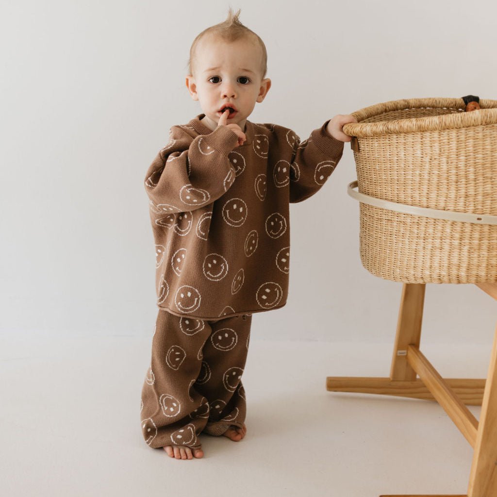 Baby in a brown patterned outfit standing next to a wicker basket on a white background