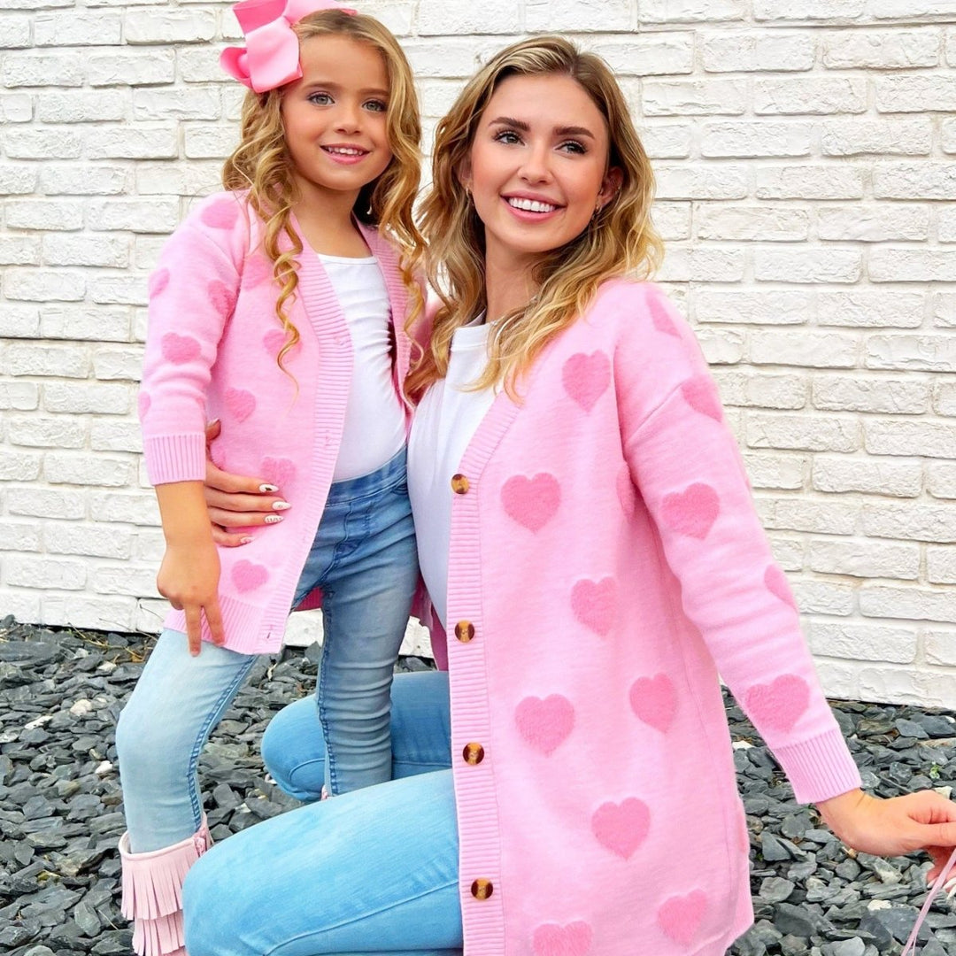 Woman and young girl wearing matching pink cardigans with heart patterns against a white brick wall.