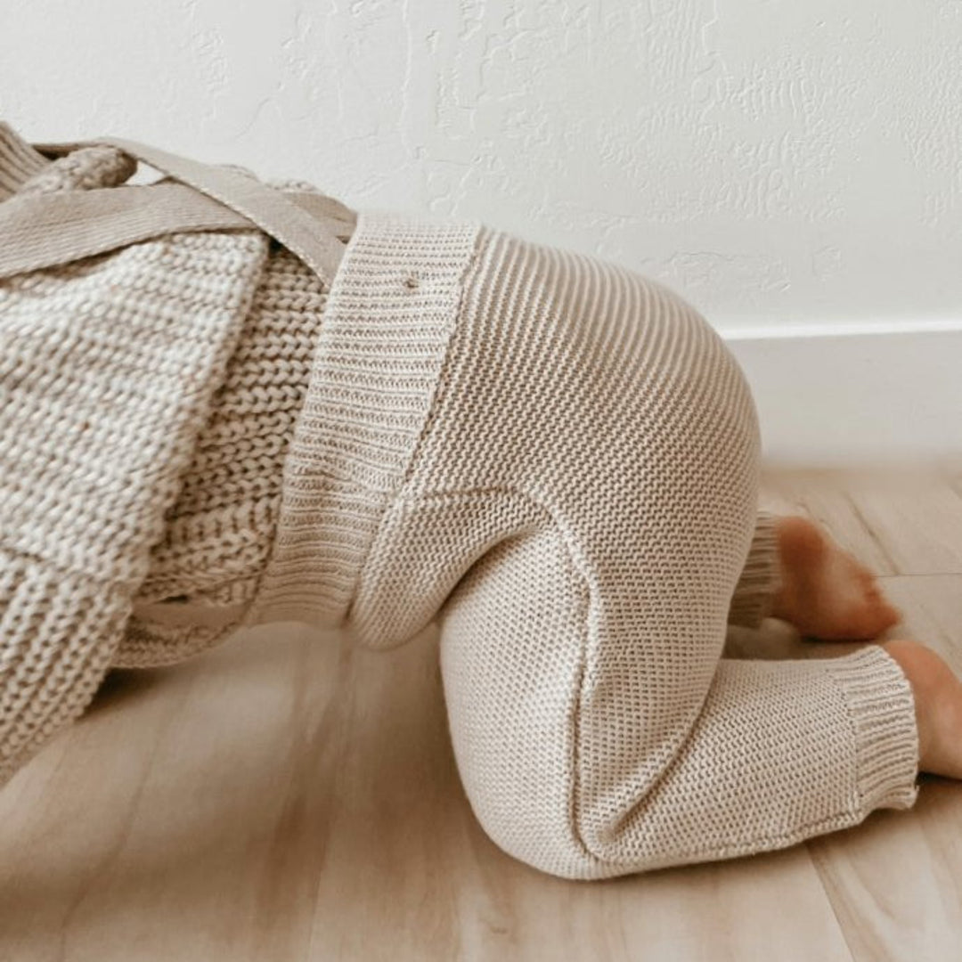 Baby wearing a beige knit sweater and pants lying on a wooden floor.