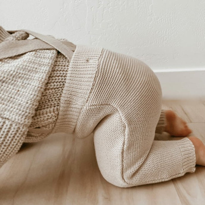 Baby wearing a beige knit sweater and pants lying on a wooden floor.