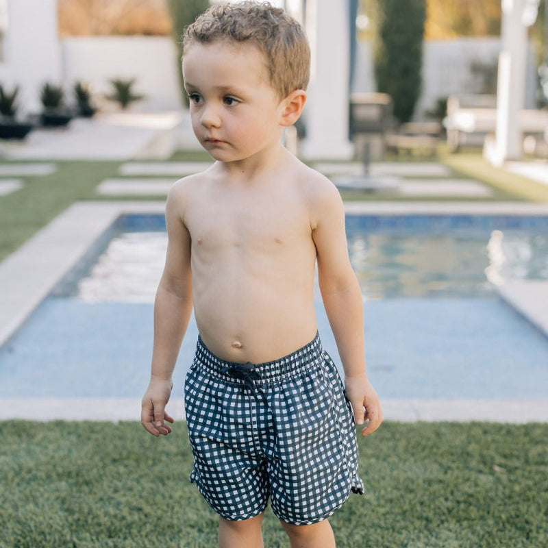 Child wearing polka dot swim shorts standing on grass near a pool.