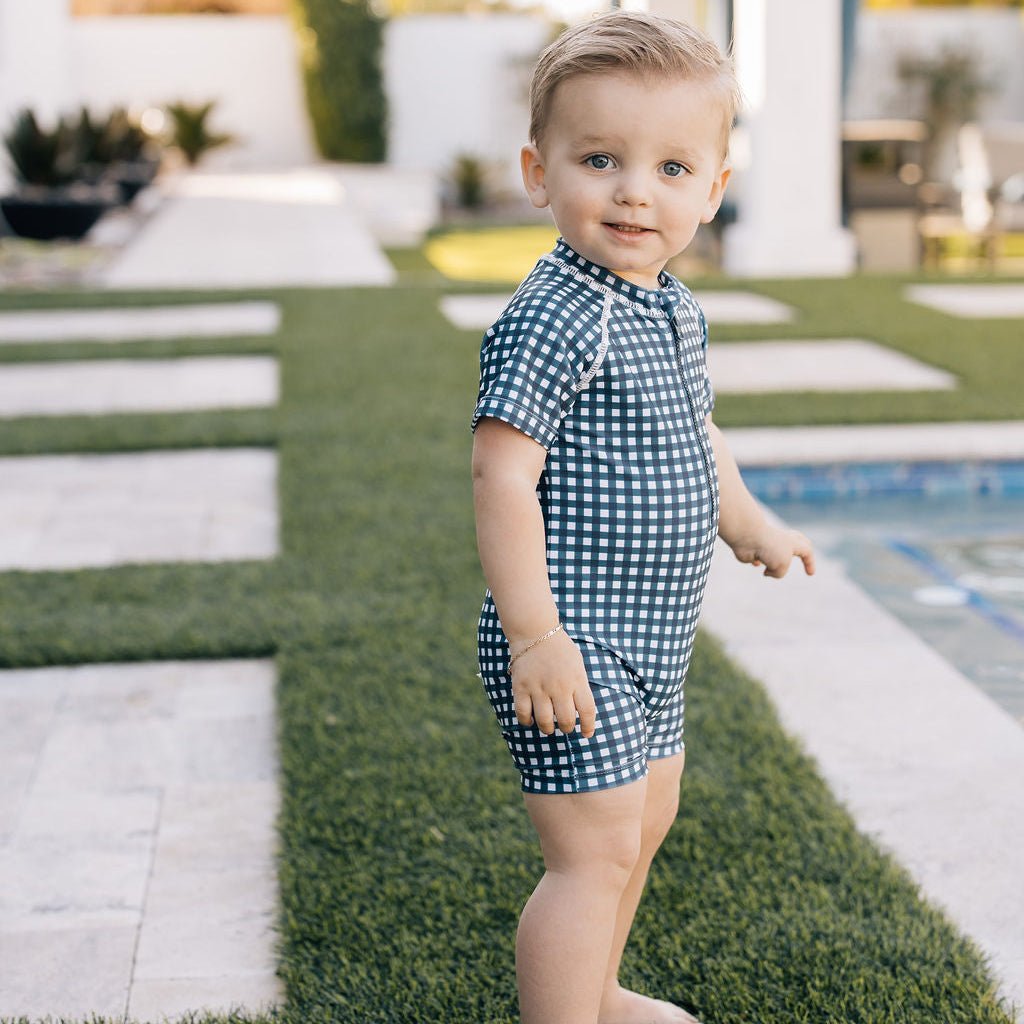 Child in a blue checkered outfit standing on grass near a pool.