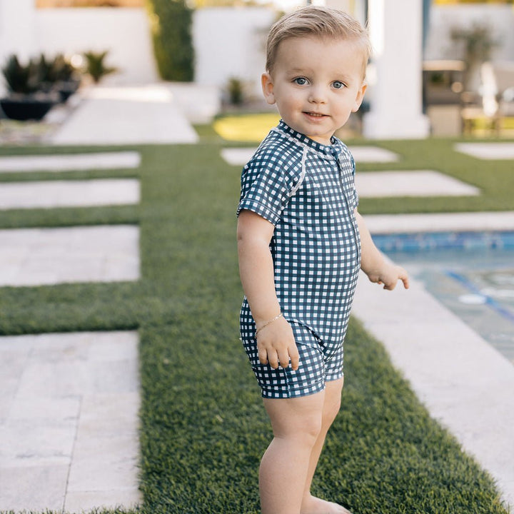 Child in a blue checkered outfit standing on grass near a pool.