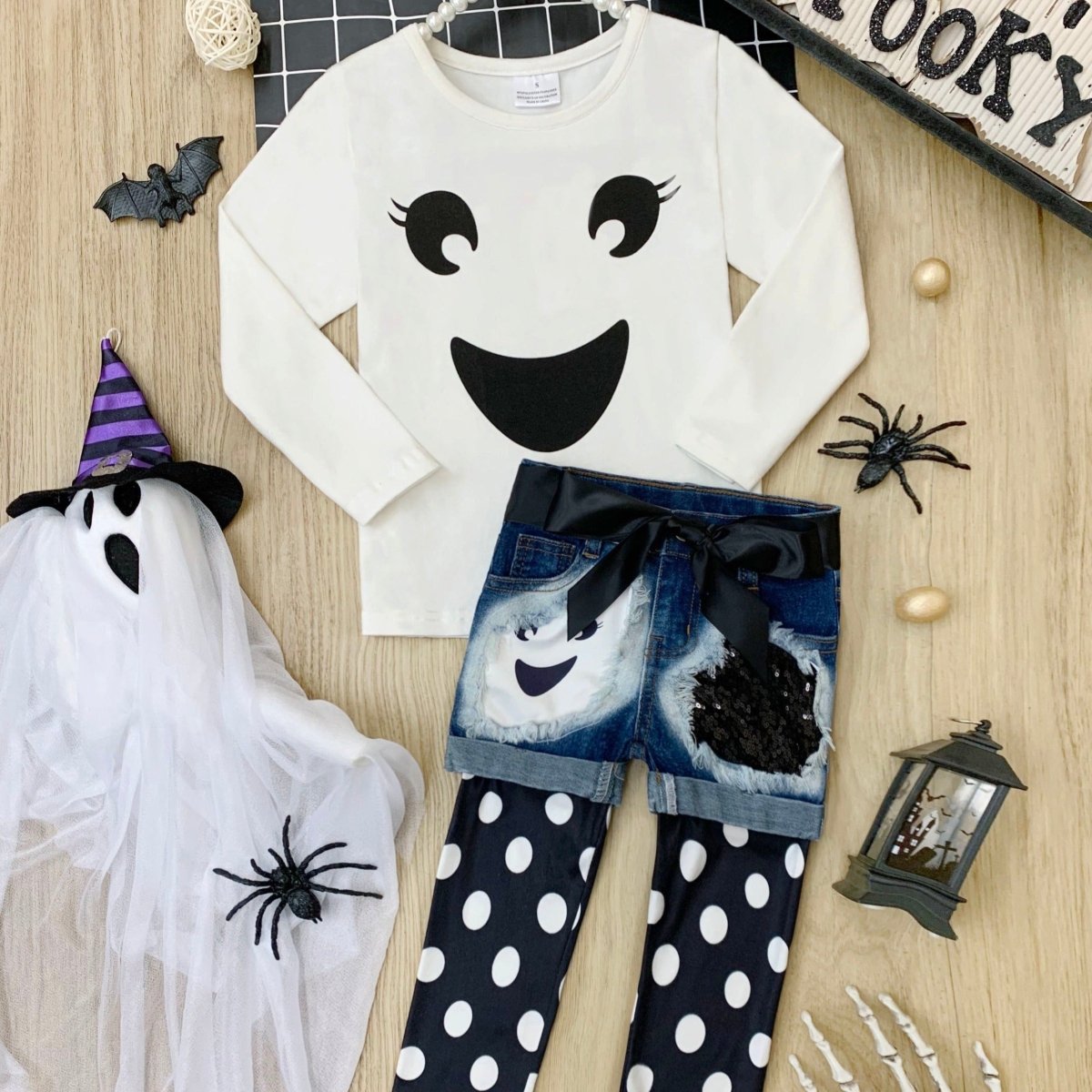 Children's Halloween outfit with ghost-themed shirt, shorts, and polka dot leggings on a wooden surface.