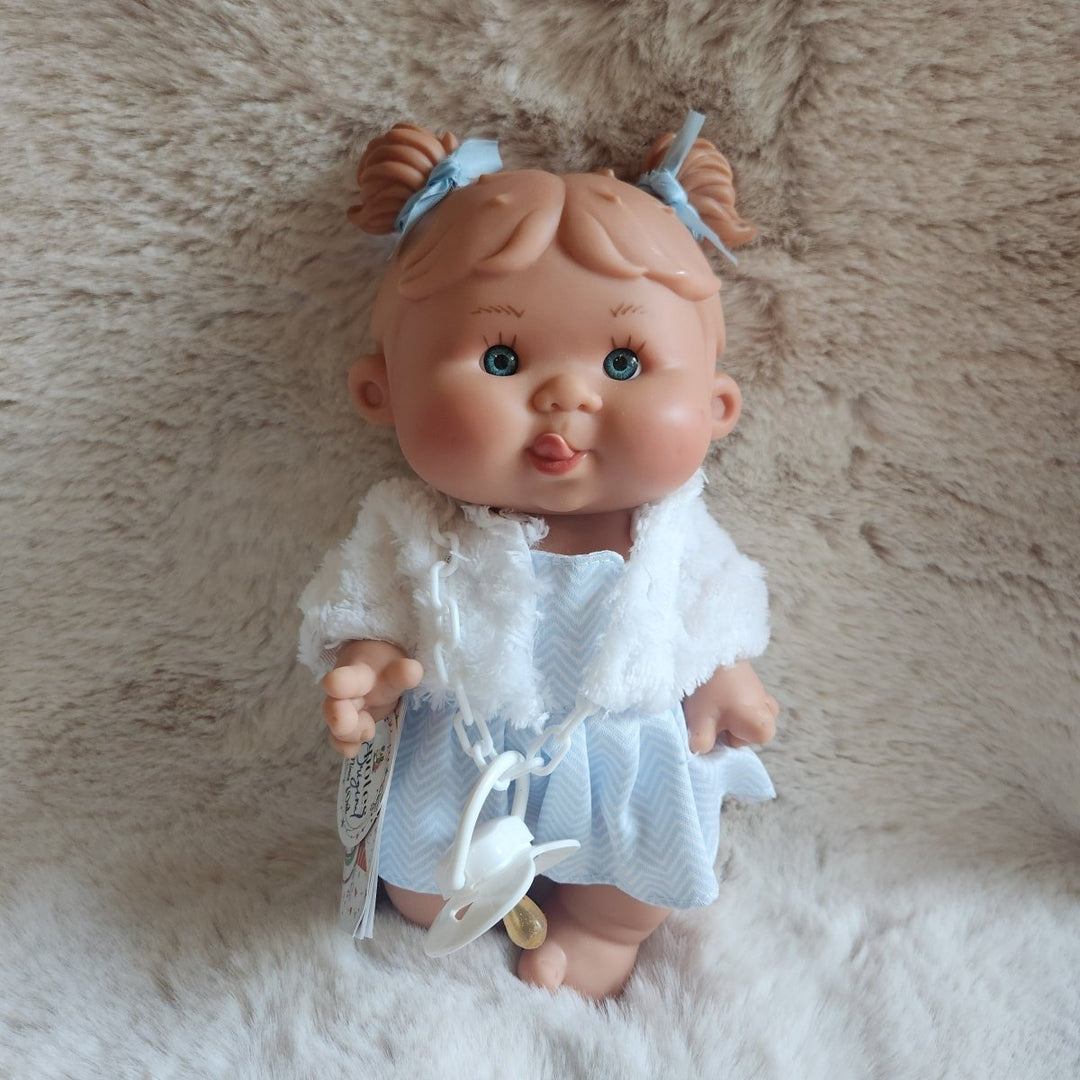 A realistic baby doll with brown hair in pigtails, blue eyes, and a pink mouth, wearing a white frilly top and light blue skirt, lying on a fluffy beige and white rug.