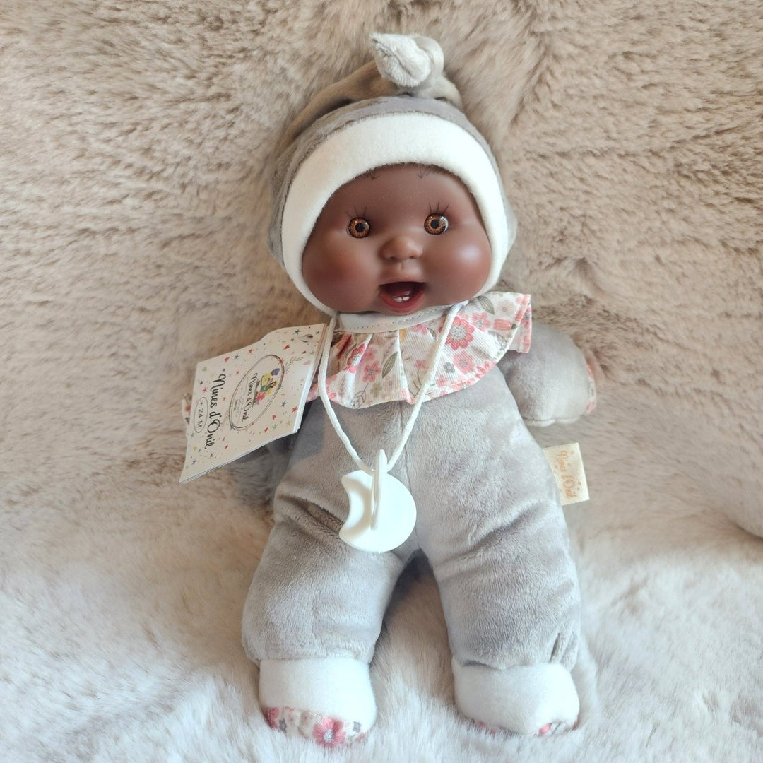 A baby doll with dark skin and brown eyes, wearing a gray and white knit hat and a light gray outfit with a pink patterned bib, lying on a beige faux fur background. 