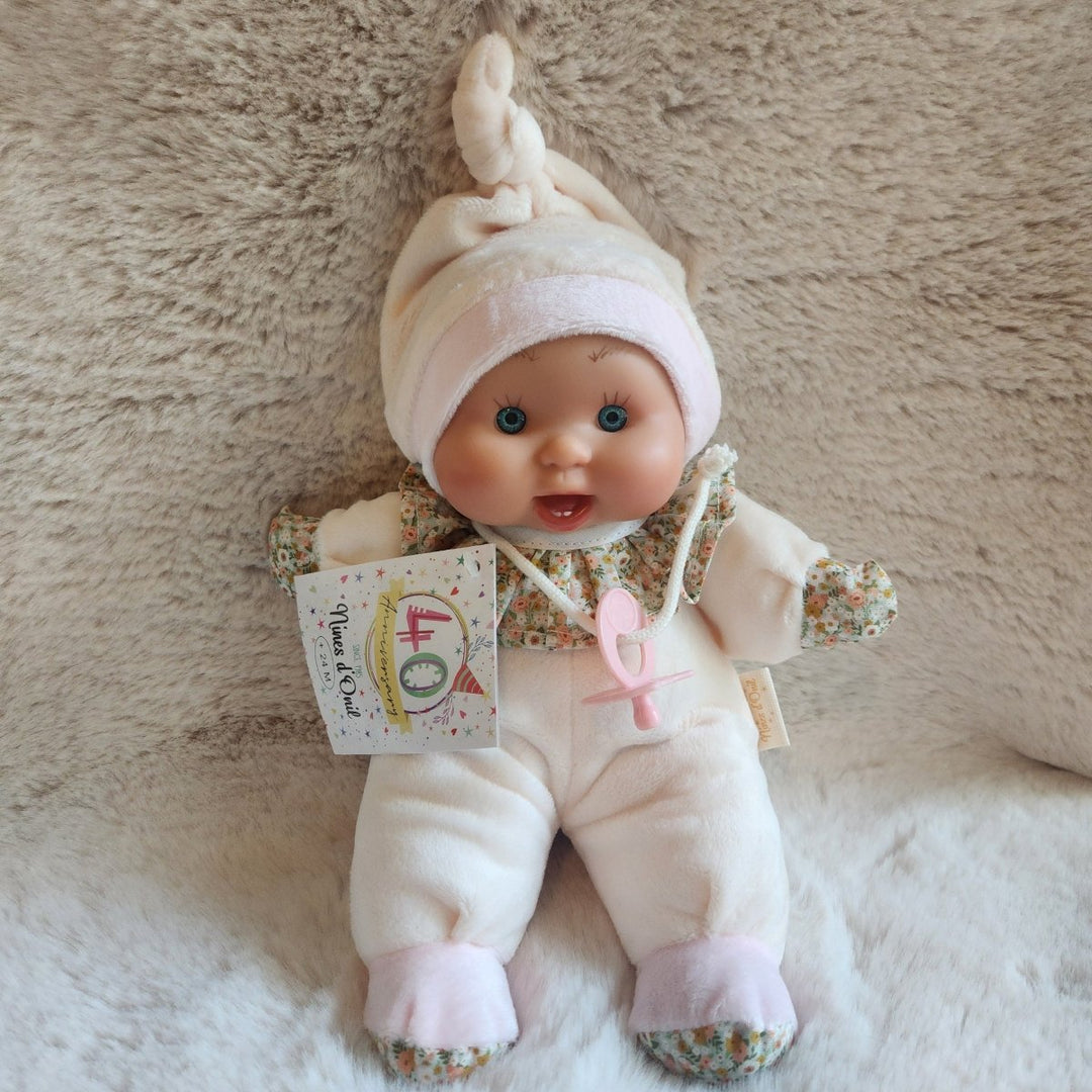 A baby doll dressed in a cream-colored onesie and matching hat, lying on a soft beige faux fur surface. The doll has blue eyes and a pacifier clip, with a small hangtag attached.