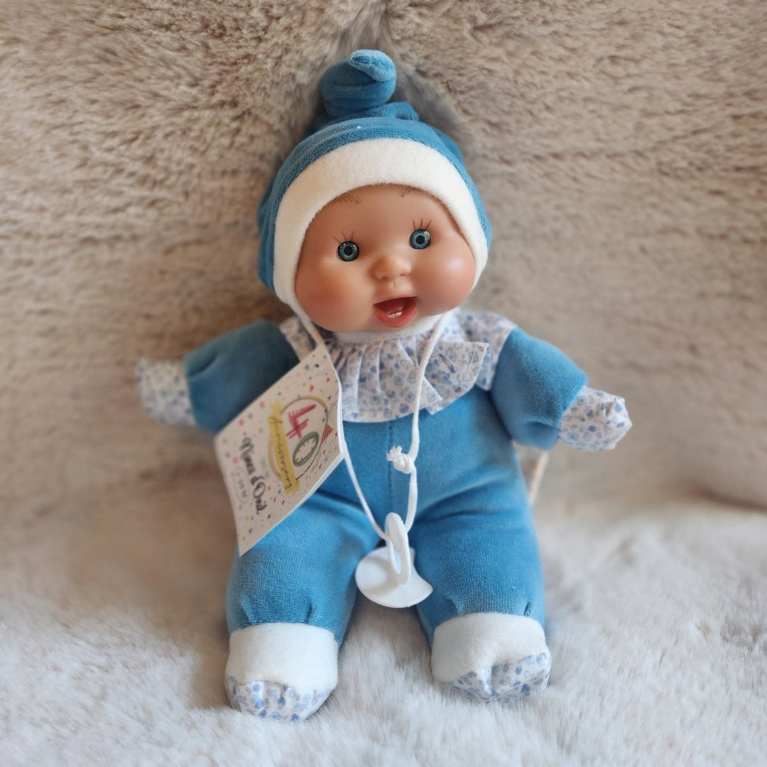 A baby doll with a blue and white outfit, blue hat, and attached product tag, posed on a soft beige textured surface.