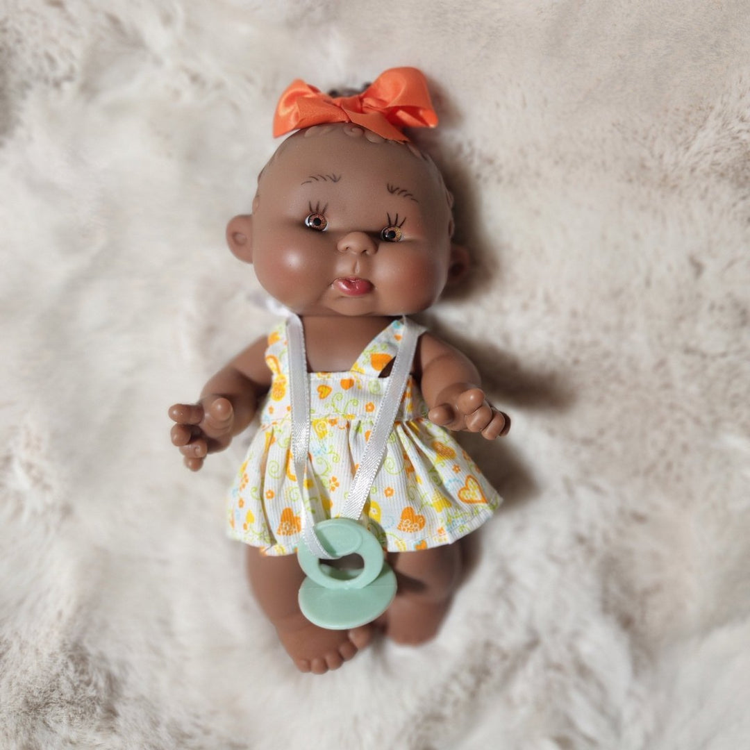 A realistic baby doll lying on a white faux fur blanket, wearing a yellow and orange floral romper with white straps and an orange bow, with a mint green pacifier in its mouth.