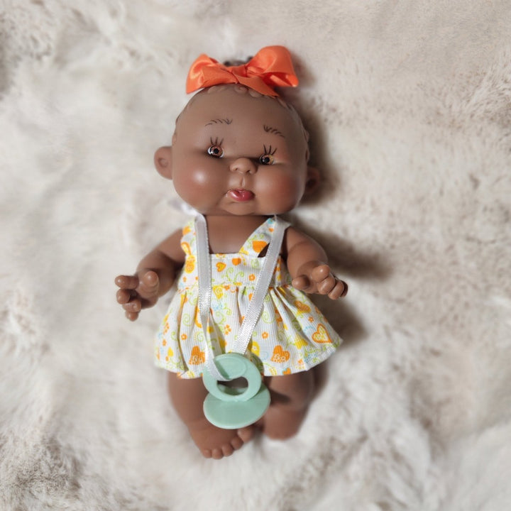 A realistic baby doll lying on a white faux fur blanket, wearing a yellow and orange floral romper with white straps and an orange bow, with a mint green pacifier in its mouth.