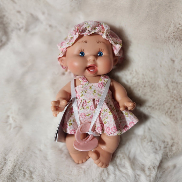 A baby doll lying on a white faux fur surface, wearing a pink floral romper and matching bonnet with a pacifier attached.