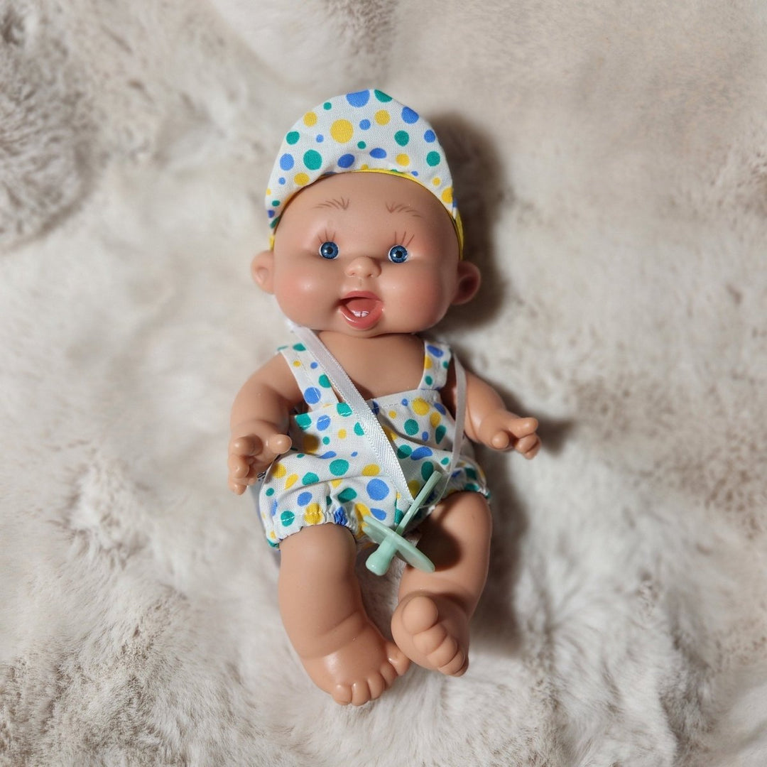 A realistic baby doll lying on a fluffy white faux fur rug, wearing a white one-piece outfit and matching bib with blue and green polka dots.