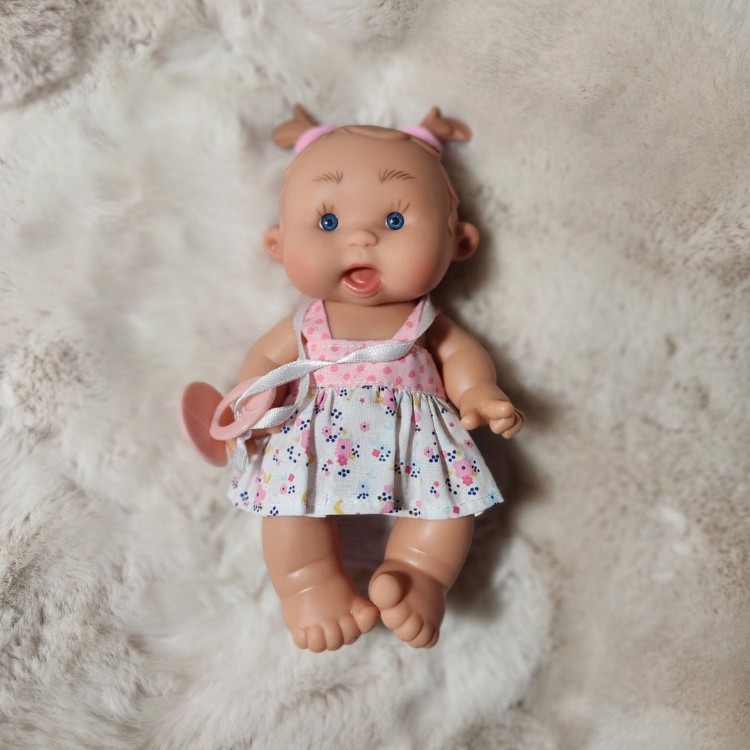 A baby doll lying down on a fluffy white faux fur surface, wearing a white dress with a pink and blue floral pattern and a pink bow tie.