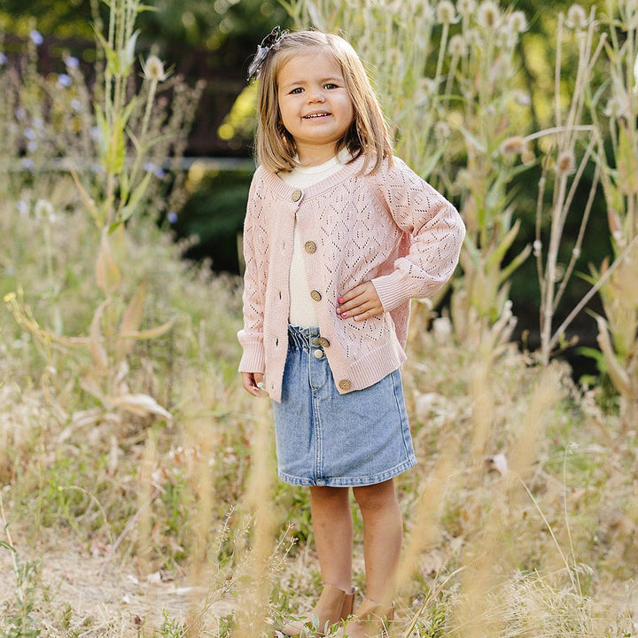 Young girl in a pink cardigan and denim skirt standing in a field of tall grass and wildflowers.